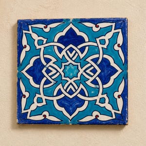 May include: A square ceramic tile featuring a symmetrical geometric pattern in shades of deep navy blue, bright turquoise, and white. The design consists of interlocking lines and floral-inspired shapes centered around an eight-pointed star.