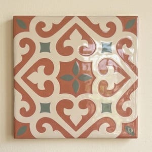 May include: A square ceramic tile featuring a symmetrical pattern of terracotta-colored scrolls and cream-colored shapes. The design includes small, sage green diamond and leaf-shaped accents arranged in a repeating geometric layout.