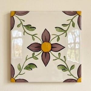 May include: A square ceramic tile with a glossy off-white background featuring a central four-petaled purple flower with a yellow centre. Green vines with leaves extend from the centre towards the corners, ending in partial purple flower petals.