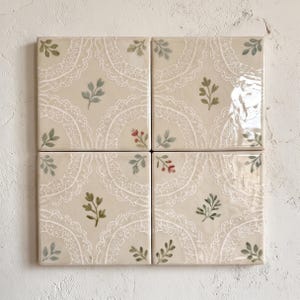 Puede incluir: Cuatro azulejos de cerámica cuadrados dispuestos en cuadrícula, con un fondo color crema y un patrón de encaje blanco. Cada azulejo está decorado con pequeñas ramas de hojas verdes pintadas a mano y diminutos capullos rojos en las esquinas.