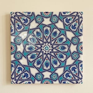 May include: A square ceramic tile featuring a complex geometric pattern with interlocking dark blue lines on a cream background. The design incorporates stylized blue and turquoise tulip motifs and small white floral accents arranged in a radial symmetry.