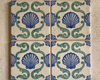 May include: A set of four square ceramic tiles arranged in a grid, each featuring a central blue seashell motif surrounded by green scrolling leaf patterns on an off-white background, with visible fine crackle glazing on the tile surfaces.