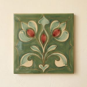 May include: A square ceramic tile featuring a symmetrical floral motif in shades of sage green, cream, and muted red. The design includes stylized petals and leaves with dark outlines, set against a solid green background with a glossy finish.