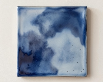 May include: A square ceramic tile featuring a glossy glaze with a blended watercolor effect in shades of deep navy blue, royal blue, and pale sky blue. The surface displays subtle dark speckles throughout the smooth, reflective finish.