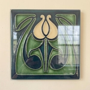 May include: A square ceramic tile featuring a raised relief design of a central cream-colored tulip-like flower surrounded by stylized green and dark teal leaves. The surface has a glossy finish with deep color variations and smooth, curved edges.