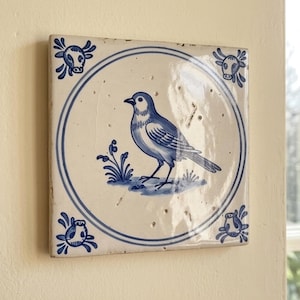 May include: A square ceramic tile with a glossy off-white finish featuring a blue hand-painted bird centered within a circular border. The four corners of the tile are decorated with small blue floral motifs, creating a classic decorative accent.