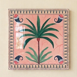May include: A square ceramic tile featuring a central green palm tree on a light pink background. The design includes four dark blue paisley motifs in the corners and a decorative border with small black dots and repeating line patterns.