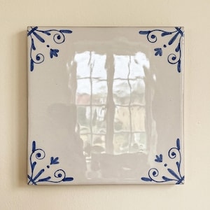 May include: A square ceramic tile with a glossy off-white finish. The tile features a hand-painted blue decorative corner motif in each of the four corners, consisting of delicate scrolls and a central petal shape, creating a symmetrical design.