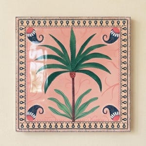 May include: A square ceramic tile featuring a central green palm tree illustration on a light pink background. The design includes four dark blue paisley motifs in the corners and a decorative black and white border with repeating swirl patterns.