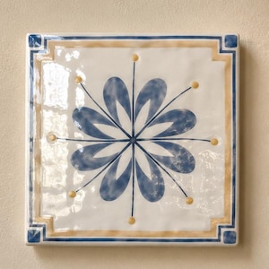 Puede incluir: Un azulejo de cerámica cuadrado con acabado blanco brillante que presenta un motivo floral azul central de ocho pétalos. Finas líneas azules se extienden desde el centro hasta pequeños puntos amarillos. El borde consta de un marco beige con cuadrados azules.