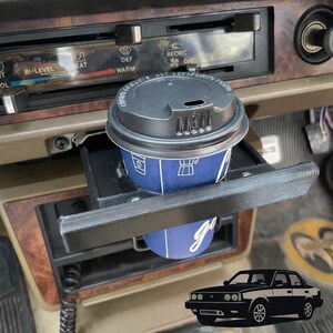 May include: A black plastic cup holder insert installed in a vehicle dashboard, holding a blue paper coffee cup with a black lid. The dashboard features wood grain trim and climate control sliders labeled with text including BI-LEVEL, DEF, and RECIRC.