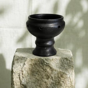 May include: A matte black ceramic bowl with a pedestal base and a series of small, evenly spaced circular holes around the upper circumference. The bowl features a smooth, rounded silhouette and a wide opening, resting on a textured stone surface.