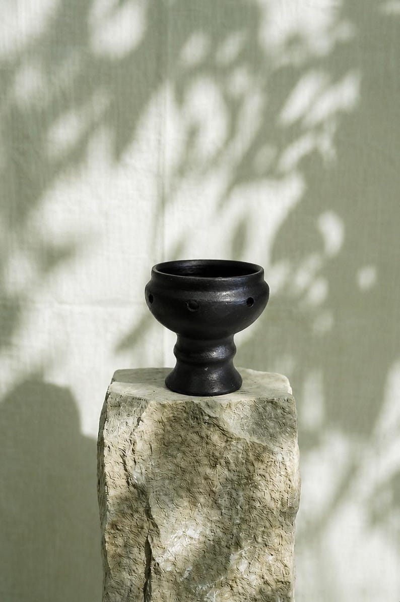 May include: A matte black ceramic bowl with a pedestal base and a series of small, evenly spaced circular holes around the upper circumference. The bowl features a smooth, rounded silhouette and a wide opening, resting on a textured stone surface.