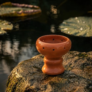 May include: A terracotta hookah bowl featuring a wide, rounded top section with a series of small, evenly spaced circular holes around the rim and a thick, flared base. The bowl has a smooth, polished finish and a warm, reddish-brown color.