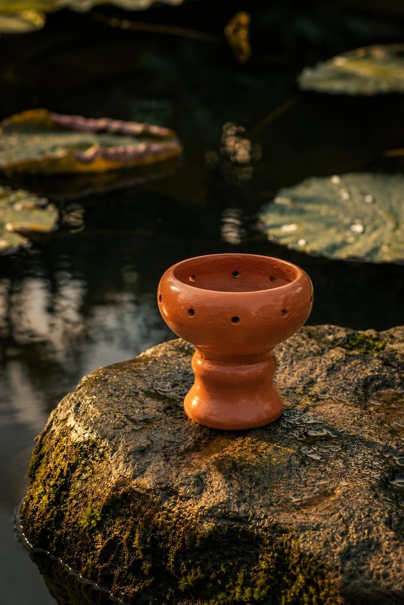 May include: A terracotta hookah bowl featuring a wide, rounded top section with a series of small, evenly spaced circular holes around the rim and a thick, flared base. The bowl has a smooth, polished finish and a warm, reddish-brown color.