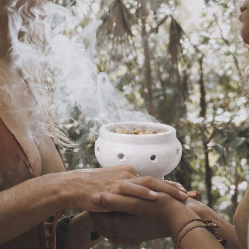 May include: A white ceramic incense burner with a rounded body and a series of small circular holes around the center. The bowl contains burning embers and aromatic materials, emitting a thick, swirling plume of white smoke into the air.