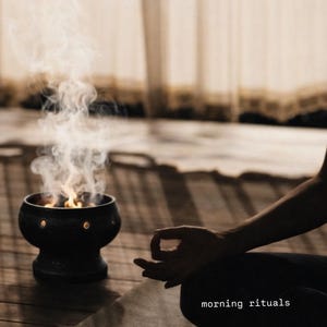 May include: A black ceramic incense burner with circular cutouts emitting smoke and flames, positioned next to a dark yoga mat. The text "morning rituals" is printed in white on the mat, which is placed on a wooden floor surface.