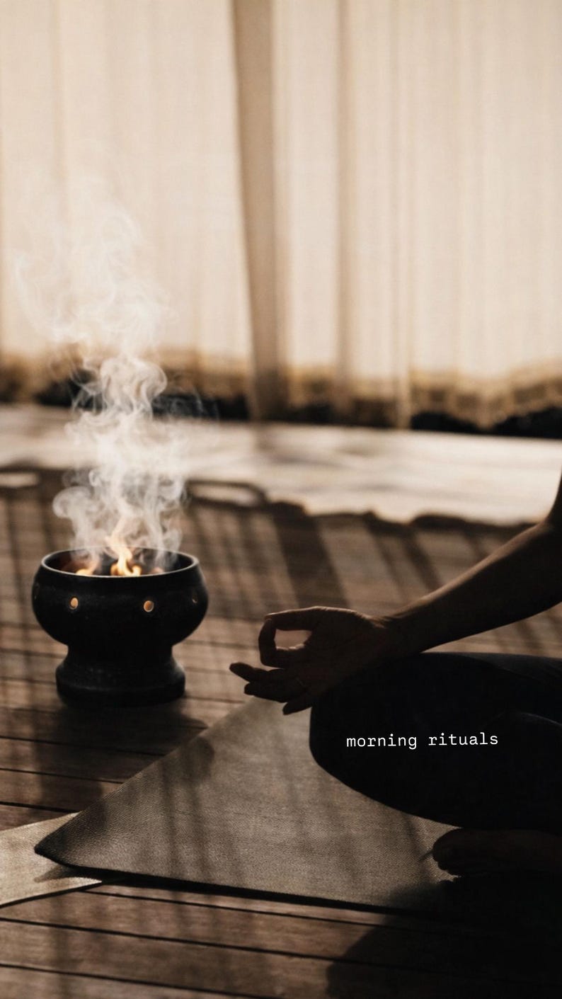 May include: A black ceramic incense burner with circular cutouts emitting smoke and flames, positioned next to a dark yoga mat. The text "morning rituals" is printed in white on the mat, which is placed on a wooden floor surface.
