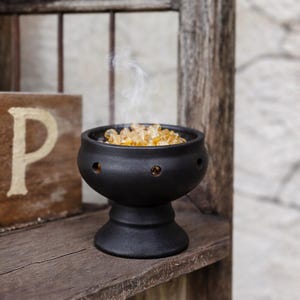 May include: A matte black ceramic bowl with a pedestal base and circular cutouts along the rim. The bowl is filled with small, translucent amber-colored resin pieces, and a thin wisp of white smoke rises from the center of the container.