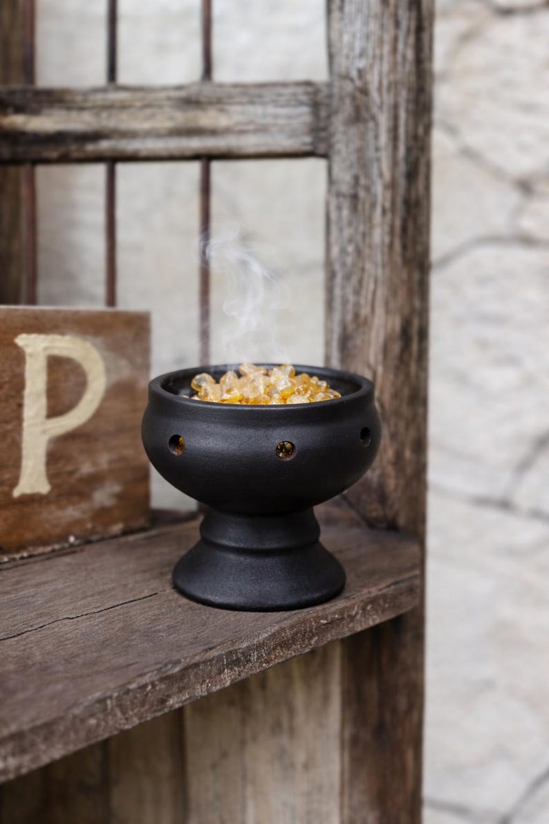 May include: A matte black ceramic bowl with a pedestal base and circular cutouts along the rim. The bowl is filled with small, translucent amber-colored resin pieces, and a thin wisp of white smoke rises from the center of the container.