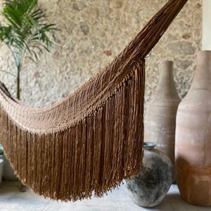 May include: A brown woven hammock with long fringe. The hammock is suspended, showcasing its texture and design. The background features a stone wall, a small palm tree, and several large, neutral-toned vases.