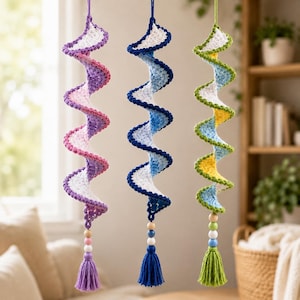 May include: Three hanging spiral decorations made of crocheted yarn. Each spiral features a white center with a different colored edge: purple, blue, and green. Each piece ends with a series of small wooden beads and a matching colored yarn tassel.