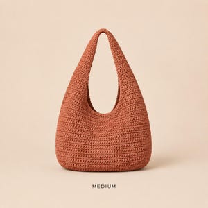 May include: A medium-sized, rust-coloured hobo bag crafted with a dense, textured crochet stitch. The bag features a single integrated shoulder strap that forms a continuous loop with the body, creating a rounded, teardrop-like silhouette.