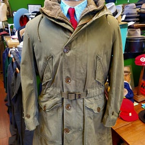 Op de afbeelding: Een olijfgroene jas in militaire stijl met een capuchon en meerdere zakken. De jas heeft een knoopsluiting aan de voorkant, een riem in de taille en een bruine met bont gevoerde capuchon. Een blauw overhemd met patroon en een rode stropdas zijn eronder zichtbaar.