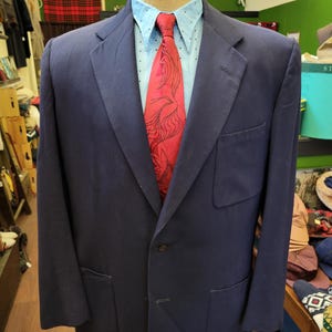 Puede incluir: Un blazer azul marino con una camisa azul claro y una corbata roja estampada. El blazer tiene dos bolsillos delanteros y un cierre de un botón. La camisa tiene un pequeño estampado de lunares.