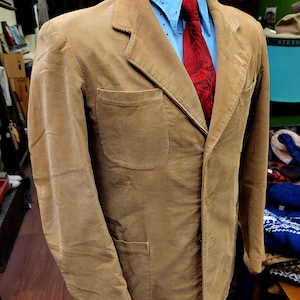 May include: A tan corduroy blazer with a notched lapel, two front pockets, and a button closure. The blazer is paired with a light blue shirt and a red patterned tie. The jacket is displayed on a mannequin.