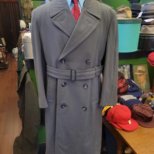 Può includere: Un trench coat grigio doppiopetto con cintura, esposto su un manichino. Il cappotto ha un colletto a lancia e dettagli con bottoni. Sotto sono visibili una camicia azzurra e una cravatta rossa. Un capo di abbigliamento esterno classico.