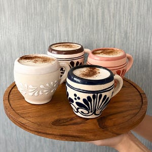 May include: Four ceramic mugs filled with a creamy beverage topped with cinnamon powder, arranged on a round wooden tray. The mugs feature distinct hand-painted patterns in white, dark blue, brown, and pink, each with a sturdy side handle.