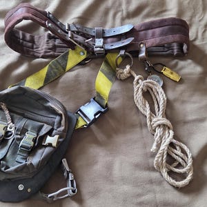 May include: A brown utility belt featuring a dark brown padded waist strap, yellow and black hazard-striped webbing, and a dark brown canvas pouch with two front pockets and plastic buckles. A coiled beige rope hangs from a metal ring on the side.