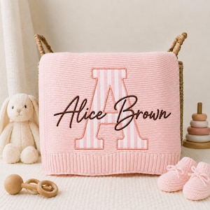May include: A soft pink knitted baby blanket featuring a large, white and pink striped letter A embroidered in the center. The name Alice Brown is stitched over the letter in a dark brown cursive font. The blanket is draped over a woven basket.