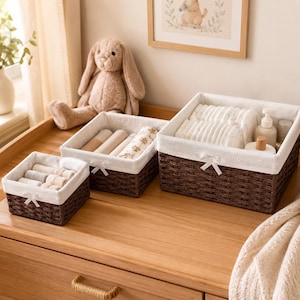 Peut inclure: Trois paniers de rangement rectangulaires en osier brun foncé avec des doublures en tissu blanc. Chaque panier présente un petit nœud en ruban blanc sur le devant. Ils contiennent des textiles blancs pliés, des articles de bébé et un flacon.