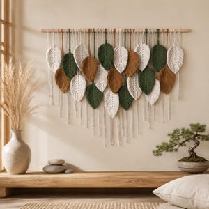 Può includere: Un arazzo in macramè con molteplici elementi in fibra a forma di foglia nelle tonalità crema, verde bosco e marrone terracotta, sospesi a un bastone di legno orizzontale con lunghi e sottili cordini verticali verso il basso.