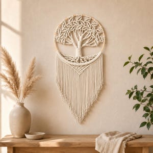 Peut inclure: Une décoration murale circulaire en corde macramé blanc cassé représentant un arbre dans un cercle en bois. L'arbre possède un tronc central et des branches aux nœuds en forme de feuilles, avec une longue frange en V suspendue en bas.