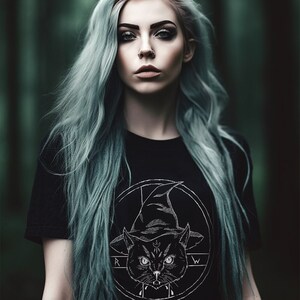 Puede incluir: Una mujer con el pelo largo azul verdoso lleva una camiseta negra con un gráfico blanco de un gato con un sombrero de bruja. El gato está rodeado de un círculo con las letras "R" y "W" a cada lado.