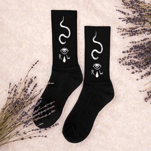 May include: A pair of black crew socks featuring a white eye, teardrop, and snake design. The socks also have the letters "R.I.W" printed on them.