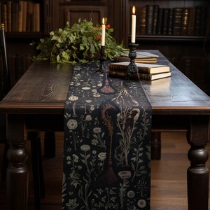Puede incluir: Una mesa de madera oscura con un camino de mesa negro con un diseño botánico con flores y una escoba. Dos velas encendidas en candelabros negros y libros están sobre la mesa. El fondo muestra una biblioteca.