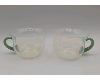 May include: Two clear glass cups with an iridescent finish and solid green handles. Each cup features a rounded bowl shape and a small circular indentation on the side, providing a distinct contrast between the translucent glass and the green.