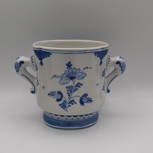 May include: A white ceramic mug featuring two handles and blue hand-painted floral patterns. The design includes a central flower motif, decorative blue accents along the rim and handles, and a solid blue band around the base with dotted details.