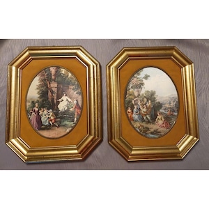 May include: Two matching octagonal wall frames with gold-toned borders and mustard-coloured inner mats. Each frame contains an oval-shaped print depicting a pastoral scene with figures in period clothing, lush greenery, stone urns, and distant landscapes.