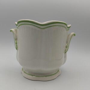 May include: A white ceramic vessel with a scalloped rim and base, featuring two side handles. The piece is decorated with thin, hand-painted green lines that follow the contours of the rim, the base, and the edges of the two side handles.
