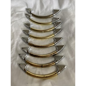May include: A set of seven curved drawer pulls arranged in a vertical column. Each pull features a central gold-toned metal bar with a smooth finish, flanked by silver-toned metal ends that have a distinct ribbed, fan-like texture on both sides.