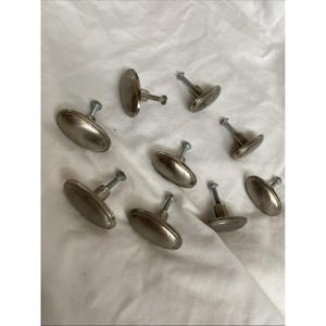 May include: A collection of eight oval-shaped cabinet knobs with a smooth, brushed silver-tone metallic finish. Each knob features a central mounting post and a threaded screw extending from the back, designed for attachment to furniture doors.