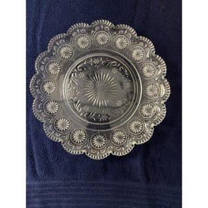 May include: A clear glass plate featuring a scalloped edge and an intricate embossed pattern. The center displays a sunburst design surrounded by floral motifs, while the border is decorated with a repeating series of circular starburst medallions.