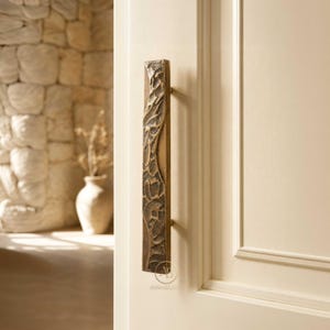 May include: A vertical rectangular door handle made of brass with a textured, uneven surface featuring deep grooves and ridges. The handle is mounted on a smooth, off-white paneled door. The metal has a warm, aged, and metallic finish.