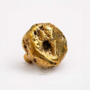 May include: A gold-colored cabinet knob with a textured, irregular, organic shape resembling a raw nugget. The surface features deep indentations and a metallic finish. The logo ASMABRASS is printed in small letters at the bottom of the object.