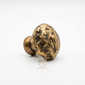 Peut inclure: Un bouton de meuble en laiton avec une surface texturée, organique et irrégulière. Le bouton présente une tête arrondie et irrégulière ainsi qu'une base cylindrique. Le métal a une finition patinée sombre. Le logo ASMABRASS est imprimé sous le bouton.